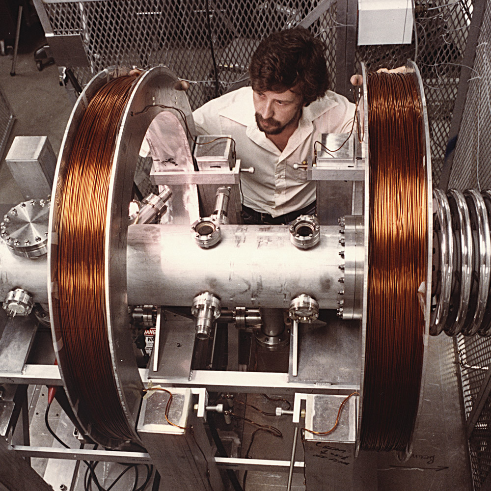 A photograph of a man between a scientific instrument that includes two spools of copper wire at either end.