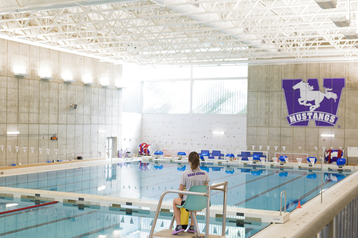 A large olympic sized swimming pool, with a lifeguarding sitting on the lifeguarding chair.