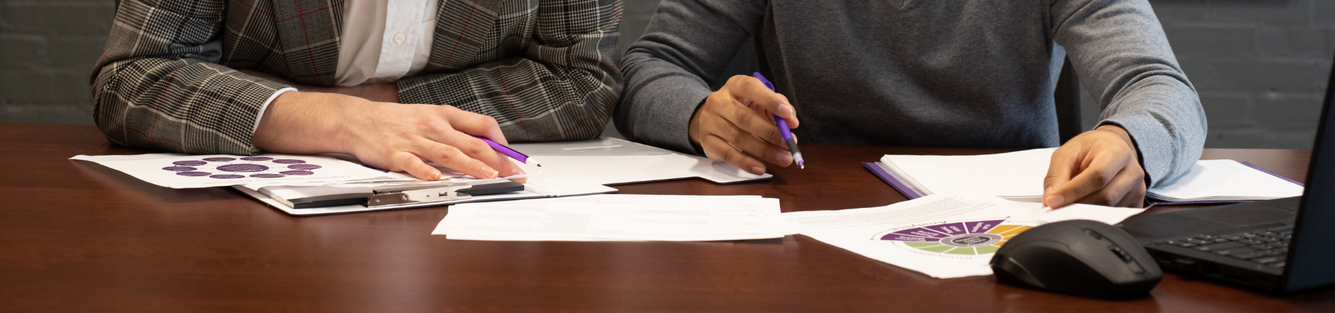 Two people sit at a table surrounded by papers, looking down at the papers with pens in their hands.
