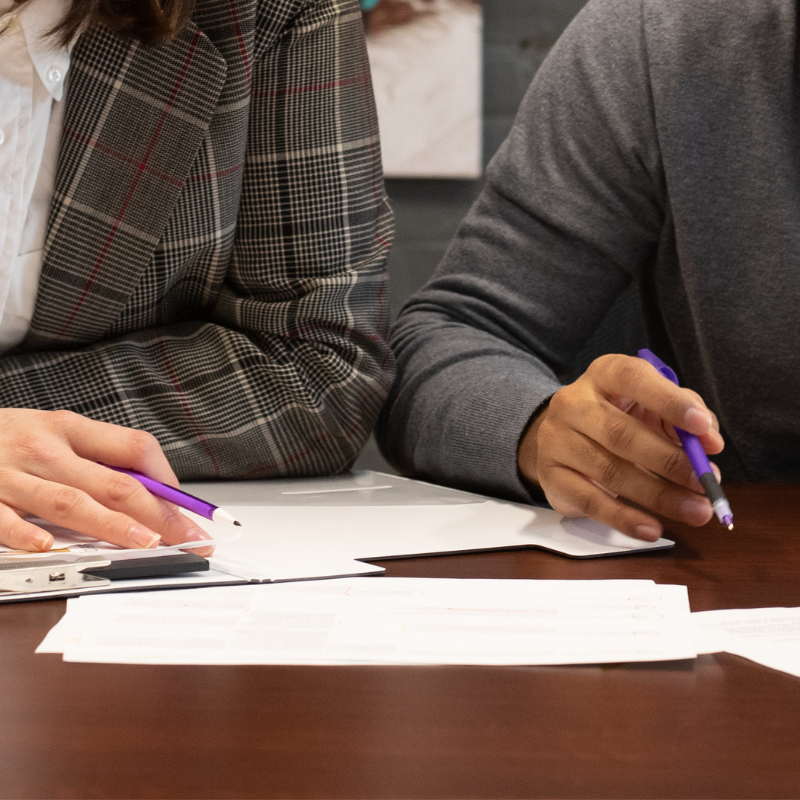 Two people sit at a table surrounded by papers, looking down at the papers with pens in their hands.