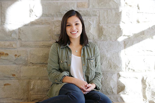 A woman is seated against a sunlit stone wall