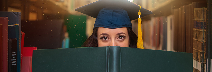 Student wearing a graduating cap and holding a book in the stacks of a library for the Banner