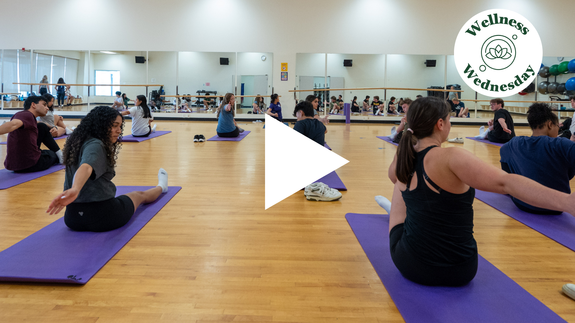Photo of Yoga Class, approx 10 participants, on the floor, doing stretches. Wood floor. Mirrors on the wall. Wellness Wednesday.