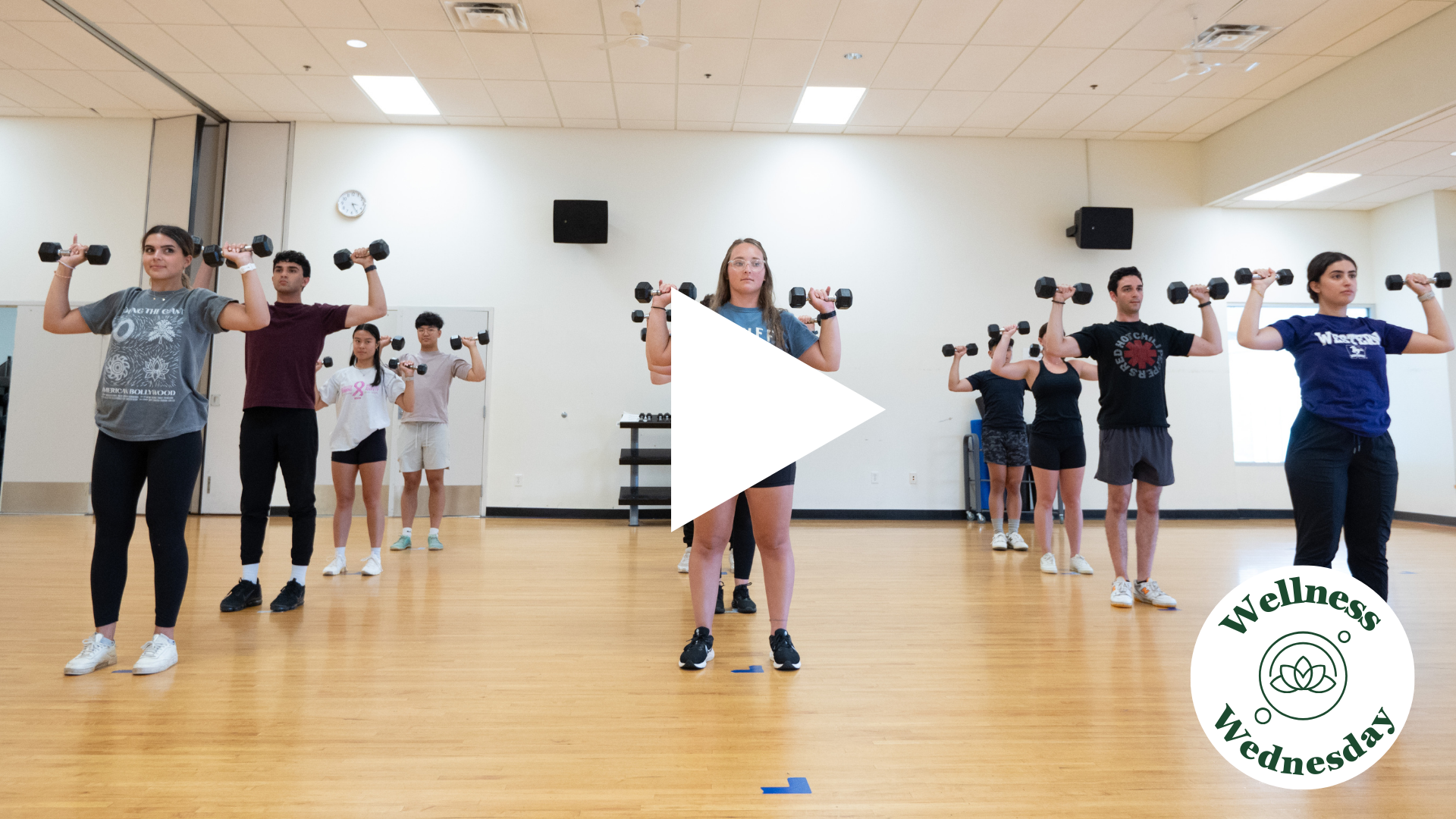 Fitness Class with weights. Gym floor. Mirrors. Multiple participants.