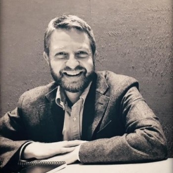 Black-and-white portrait of Endre Begby, smiling at the camera while seated at a table, wearing a blazer over a collared shirt.