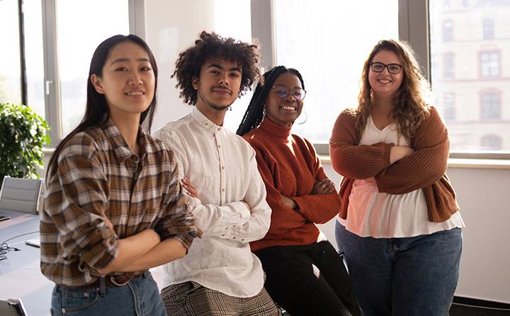 Four people standing in a row in an office setting smiling with their arms crossed.