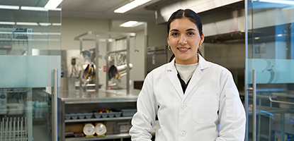 A student wears a lab coat and stands in front of doors to the commercial kitchen lab