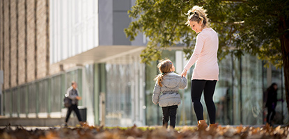 A woman and child walk outside near a building while holding hands.