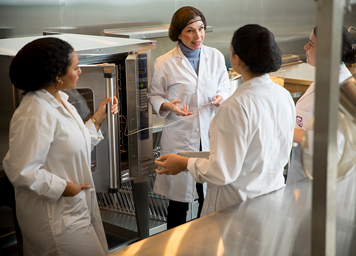 A professor speaks to students in the food lab while all are wearing lab coat and hair nets. They are standing in front of a commercial oven.