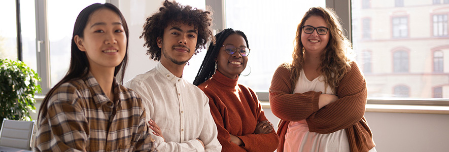 Four people standing in a row in an office setting smiling with their arms crossed.