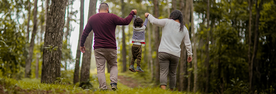 A man and woman hold a child up while walking through a wooded path on a nice day.