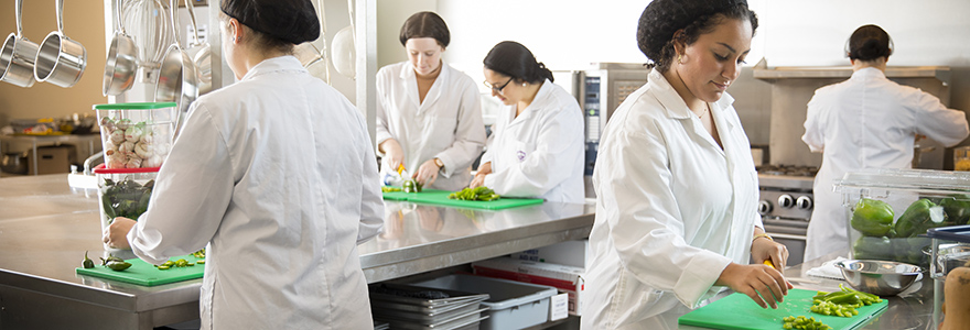 Studnets in lab coats and hair nets are working in a food lab, cutting vegetable and working on other food preparation. 