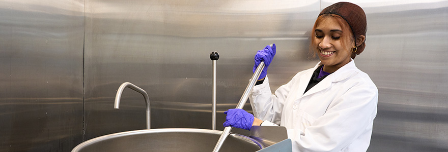A student in a lab coat stirs a giant pot with a paddle.