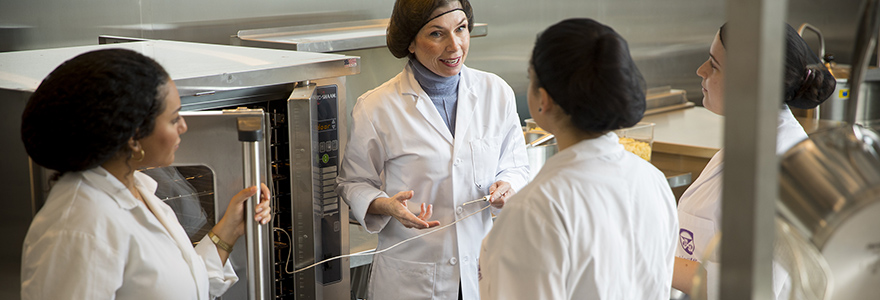 A professor speaks to students in the food lab while all are wearing lab coat and hair nets. They are standing in front of a commercial oven. 