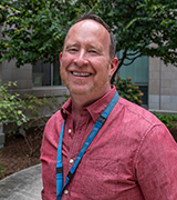 A headshot photo of Clark Heard outdoors near trees and wearing a pink button up shirt
