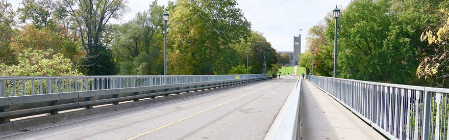 A clear sunny day. The University Drive bridge leads up to the University College tower. The campus is quiet, with just a few people in the distance.