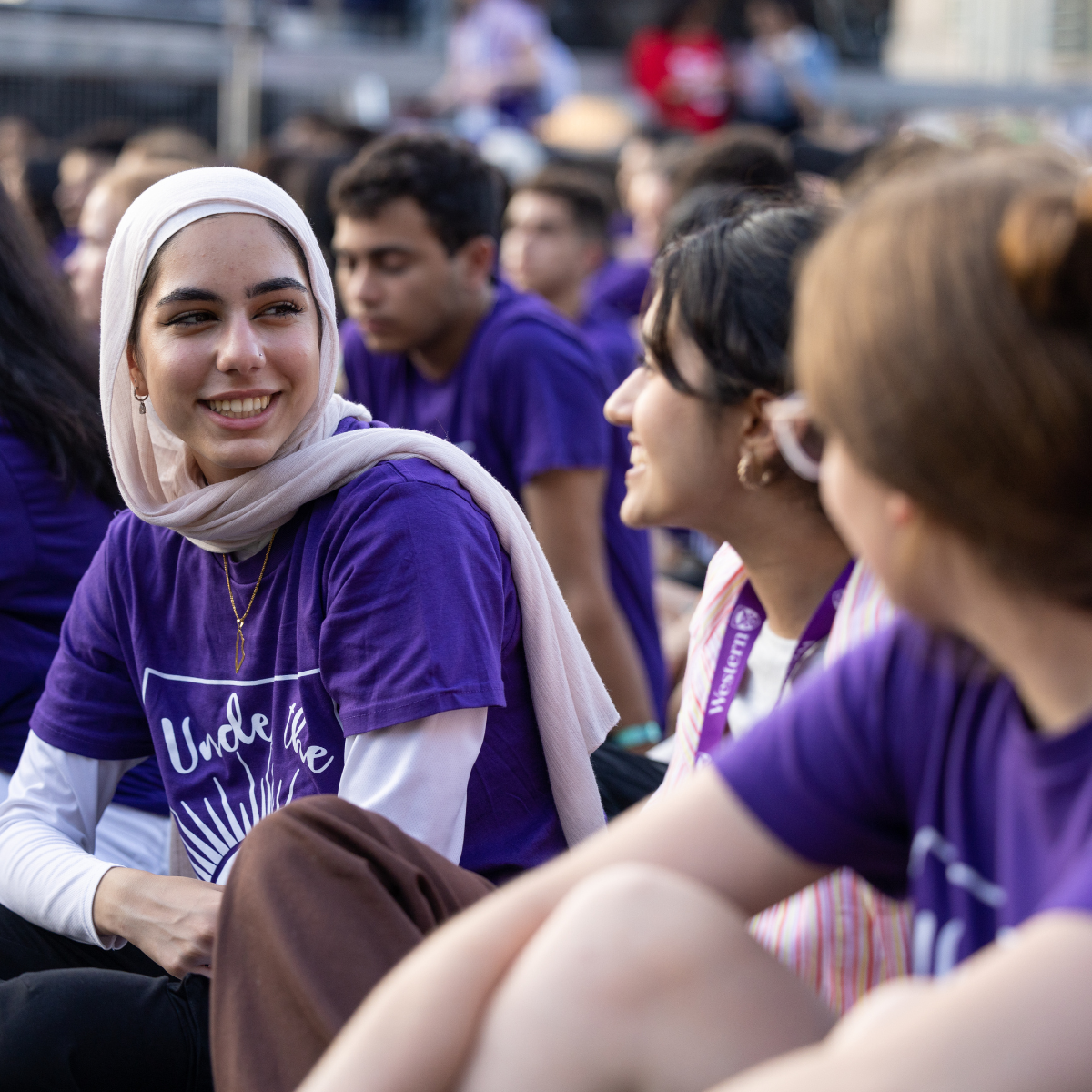 Sitting in a field amongst a diverse crowd, a girl wearing a headscarf turns towards someone off-camera