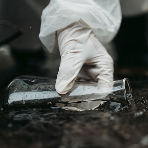 Gloved hand dips glass beaker into running water.
