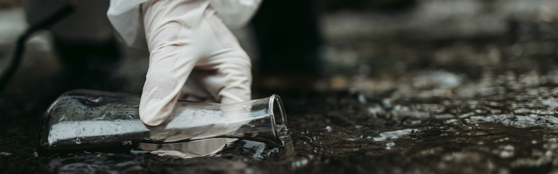 Gloved hand dips glass beaker into running water.