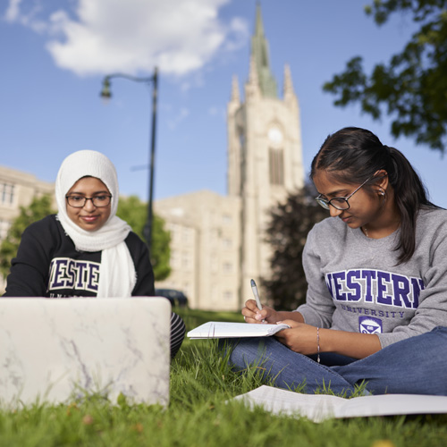 Two Western students sit on a lawn in front of Middlesex tower studying