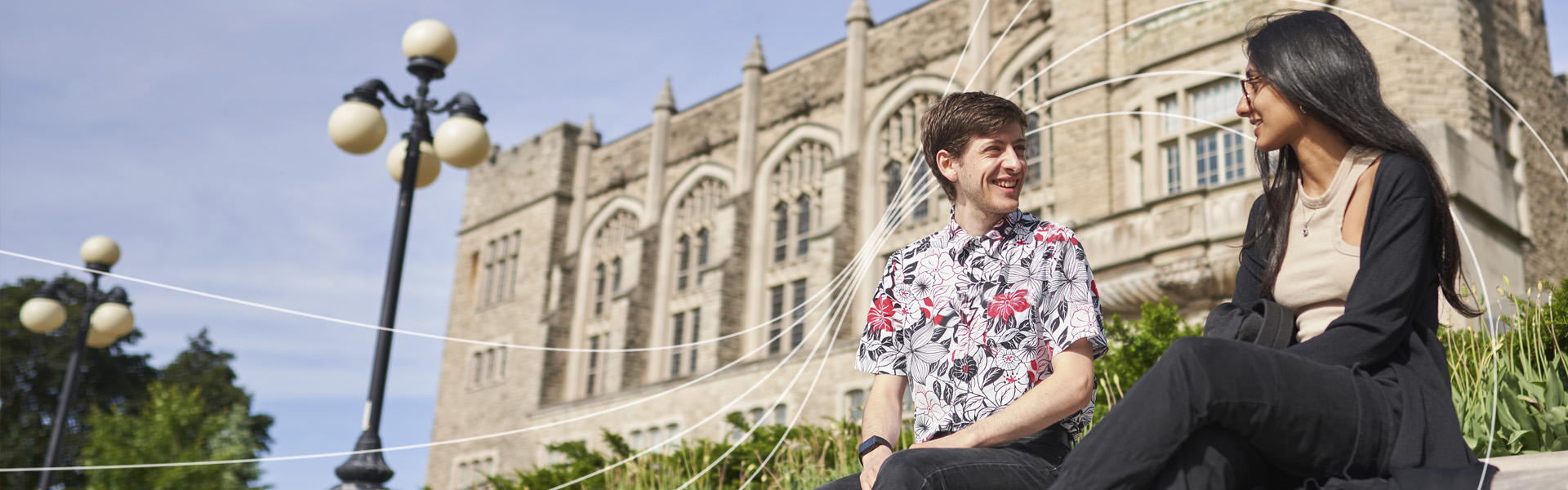 Two students sitting on campus looking to each other smiling. White wavy lines across the photo.