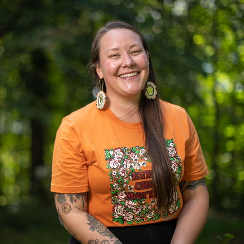 Photo of Indigenous woman smiling in front of greenery wearing an orange t-shirt
