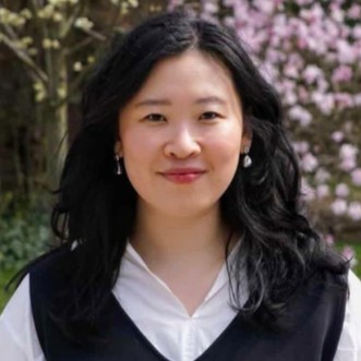 A woman with long, wavy black hair stands outdoors, smiling gently at the camera. She is wearing a white collared blouse under a dark sleeveless vest, with small dangling earrings. The background features lush greenery and soft pink blossoms, creating a warm, natural setting.