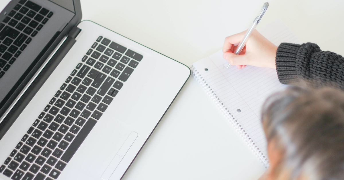 A woman with a laptop and a notebook sitting at a desk writing