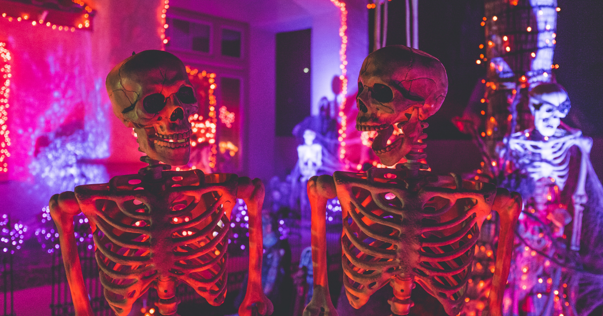 Two decorative skeletons standing near a white house with neon string lights wrapped around them