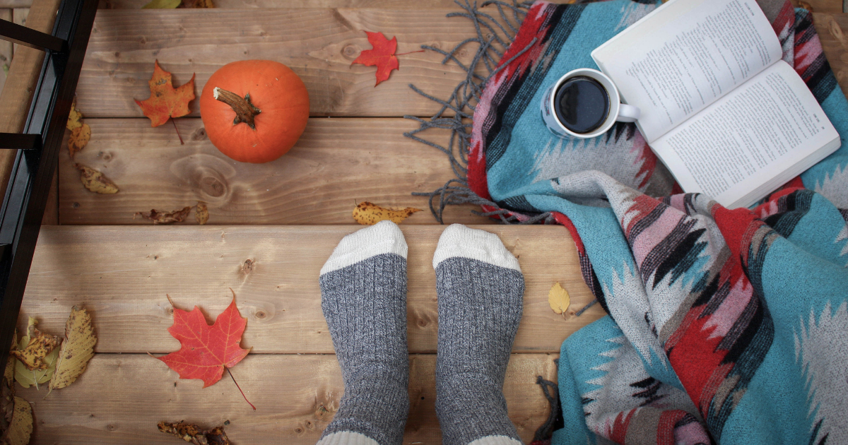 Person wearing grey socks sitting on a deck with a book surrounded by fall leaves