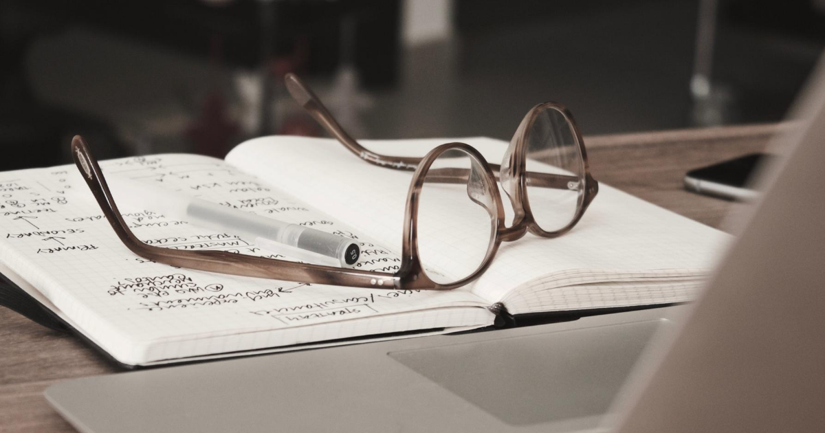 Glasses, an open notebook, and a laptop sitting on a desk