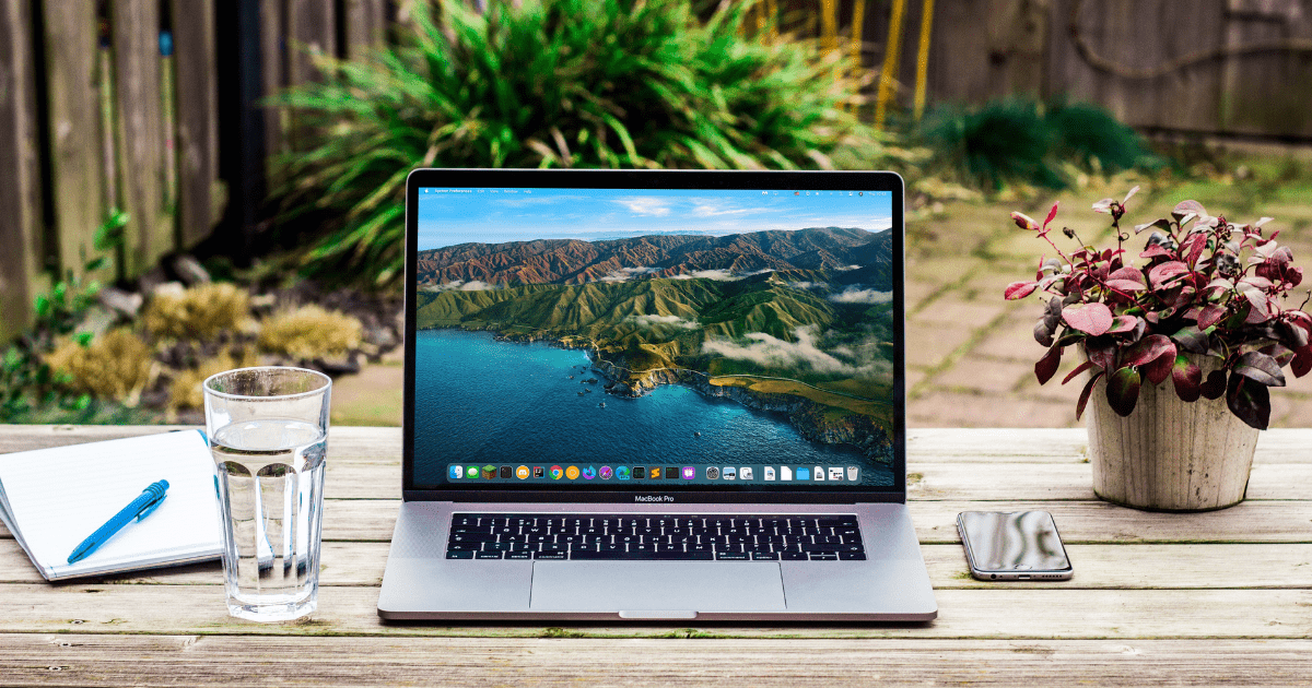 A laptop sitting on a table outside with a notebook and glass of water next to it