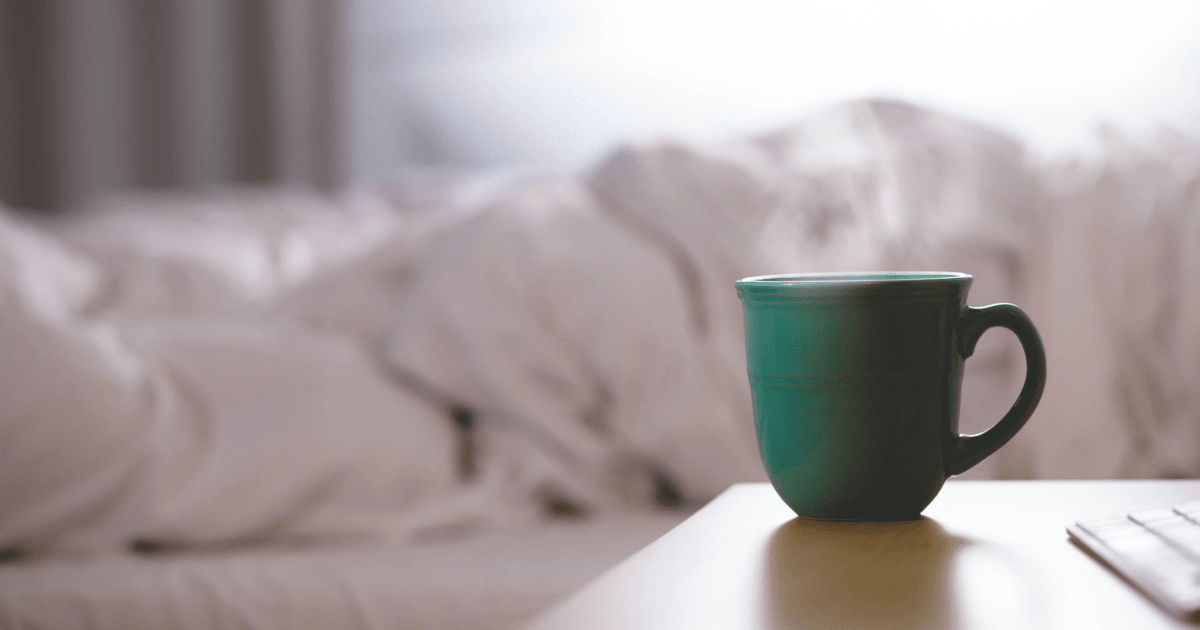 A green coffee mug with steam coming from it sitting on a table beside a bed