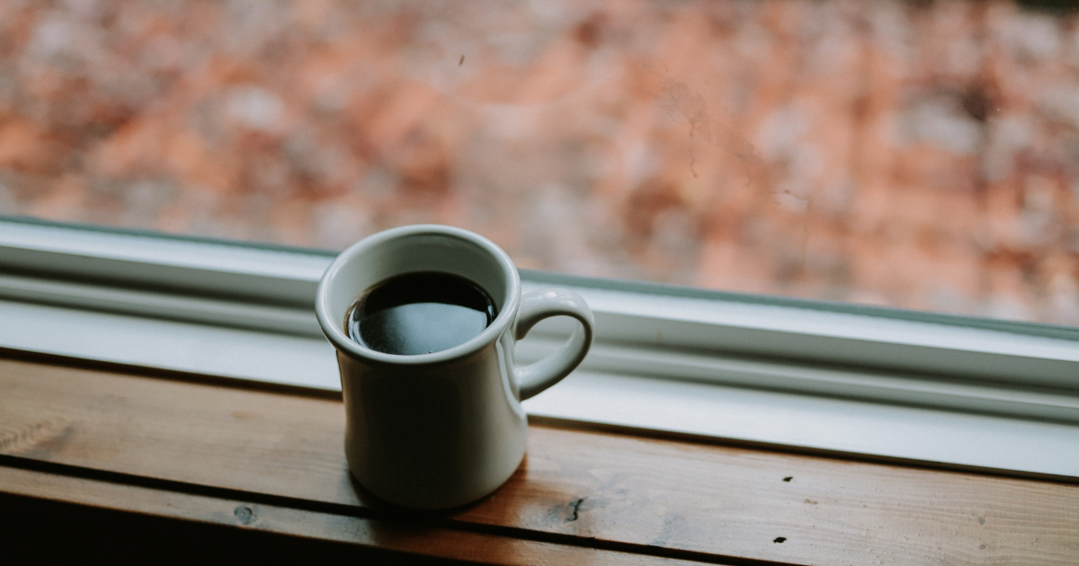 A photo of a white ceramic mug sitting on a windowsill with fall leaves in the background