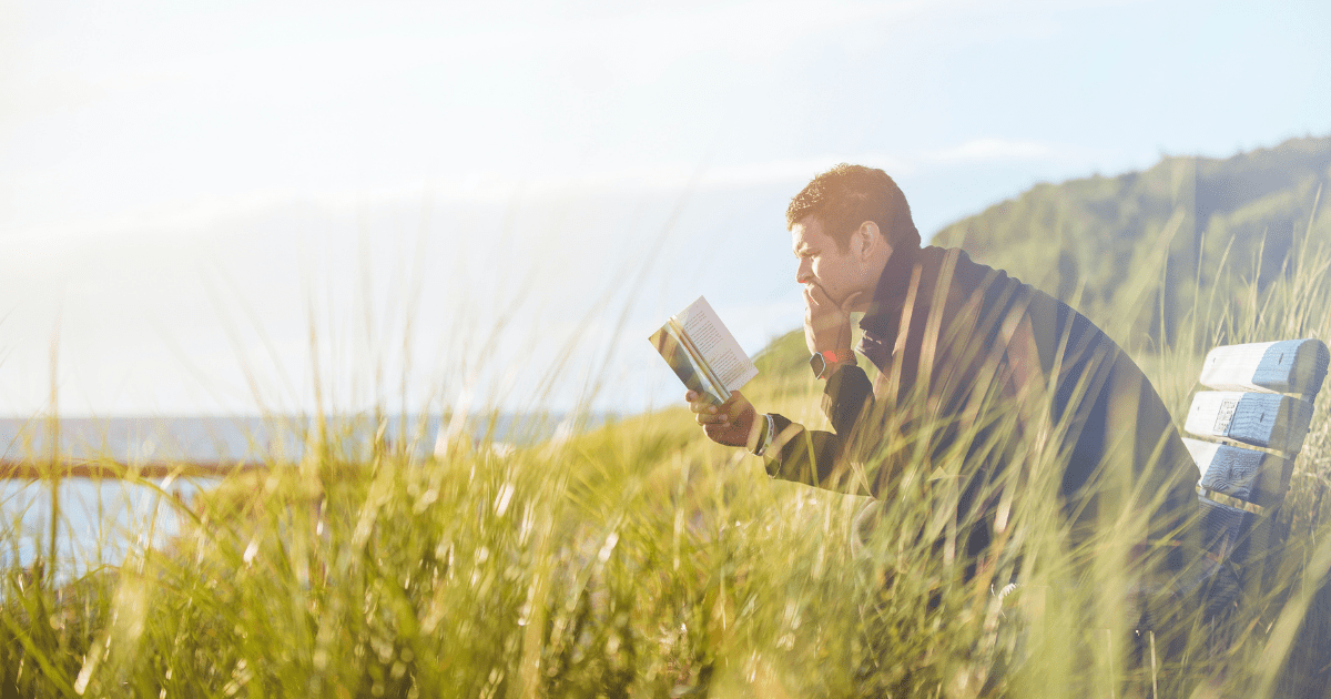 A man reading a book on a bench near a lake during the daytime