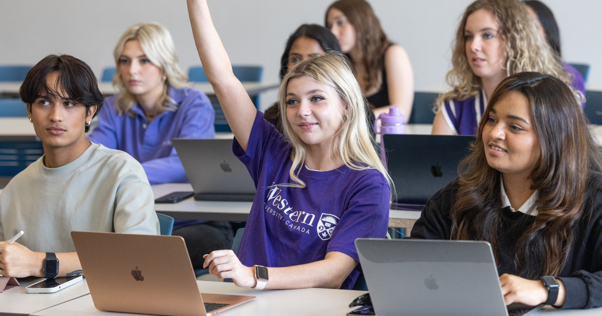 A group of students sitting in a lecture hall, one of them raising their hand.