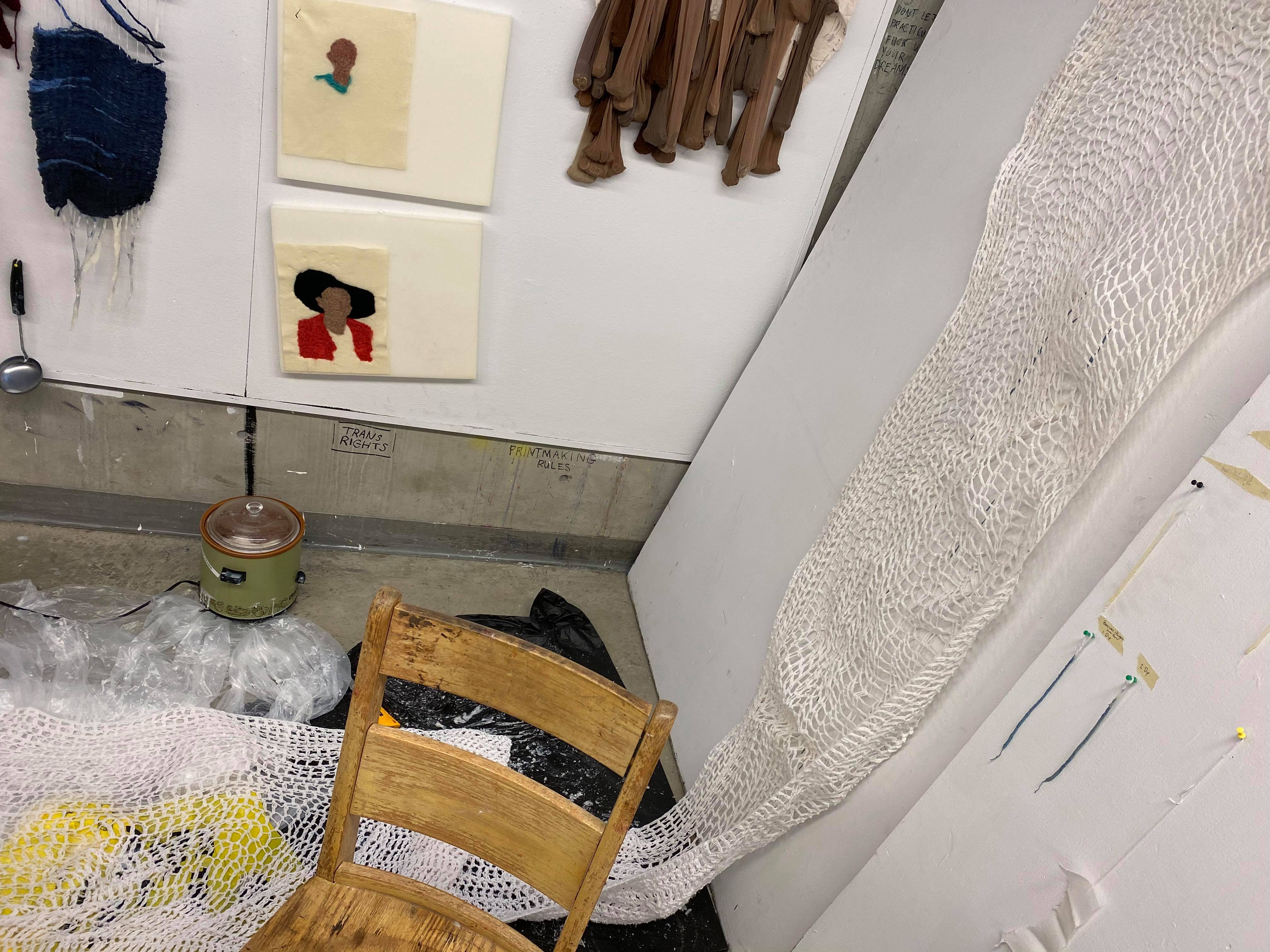 Photo of an emmpty chair, fabric, and samples of art hanging on a wall.
